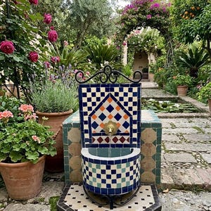 Può includere: Fontana decorativa per esterni con un design a mosaico in un rigoglioso giardino. La fontana presenta un motivo a piastrelle blu, bianche e arancioni, un rubinetto in ottone e un piccolo bacino. Il giardino comprende piante in vaso, fiori e un sentiero in pietra.