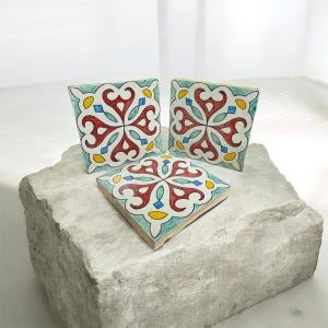 Peut inclure: Trois carreaux de céramique carrés avec un motif floral coloré. Les carreaux présentent un motif rouge, bleu, jaune et turquoise. Les carreaux sont exposés sur une surface en pierre grise claire rugueuse.