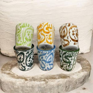 May include: Six small ceramic cups stacked in pairs. The top cups feature colorful Arabic calligraphy in green, orange, and brown. The bottom cups have matching calligraphy in dark green, blue, and light green. The cups are on a stone surface.