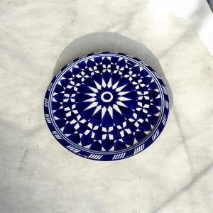 Peut inclure: Une assiette ronde en céramique ornée d'un motif géométrique bleu profond et blanc. Le design se compose d'un cercle central entouré de formes étoilées rayonnantes et de motifs floraux répétés, avec une bordure rayée sur le pourtour.