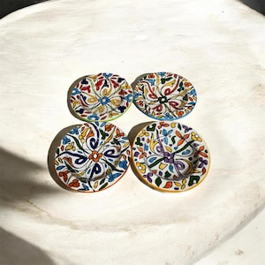 Peut inclure: Quatre petites assiettes rondes en céramique peintes à la main. Chaque assiette présente un fond blanc avec un motif floral coloré en rouge, bleu, jaune et vert. Les bords sont peints de différentes couleurs : vert, bleu, orange et jaune.