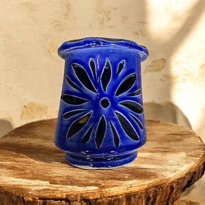 Peut inclure: Un photophore en céramique bleu cobalt avec un motif floral découpé. Le photophore cylindrique a une base et un sommet évasés. Il est posé sur une surface en bois rustique.