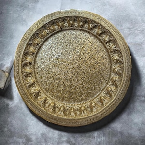 May include: A circular gold-colored metal tray featuring an intricate embossed pattern. The center displays a repeating geometric floral design, while the wide border is decorated with a series of raised, swirling leaf-like motifs.