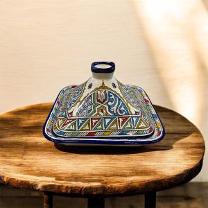 May include: A colorful, ceramic tagine pot with a lid, featuring intricate geometric and floral patterns in blue, green, red, and yellow. The pot sits on a wooden table, showcasing its decorative design.