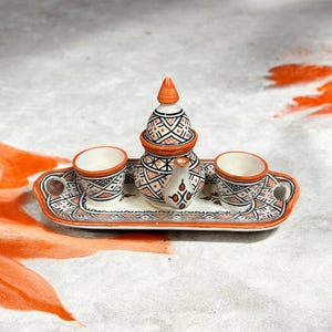 Peut inclure: Un service à thé en céramique composé d'un plateau rectangulaire à deux anses, d'une théière avec couvercle et de deux petites tasses. L'ensemble présente un motif géométrique peint à la main en noir et orange sur fond crème.