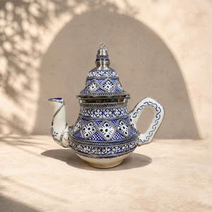 Peut inclure: Une théière en céramique avec une base blanche ornée de motifs géométriques bleus complexes et d'accents en métal argenté. La théière présente un couvercle pointu, une anse incurvée et un bec verseur, décorés de lignes et formes bleues.