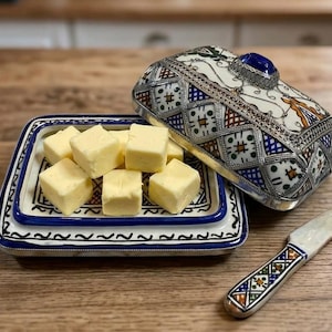Peut inclure: Un beurrier décoratif avec un couvercle argenté et bleu à motifs, un plat de service rectangulaire assorti avec des cubes de beurre et un petit couteau. Le beurrier et le couteau présentent des motifs complexes. Les cubes de beurre sont jaunes.