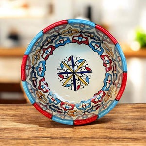 Könnte beinhalten: Dekorative Keramikschale mit einem farbenfrohen, aufwendigen Design. Der Rand ist rot und blau bemalt, das Innere weiß mit einem zentralen Blumenmuster in Gelb, Blau und Rot. Die Schale steht auf einer Holzoberfläche.