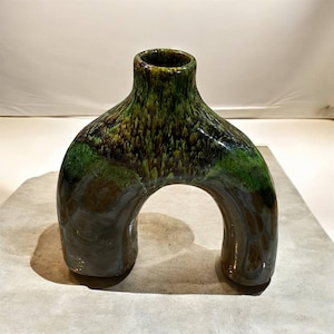 Peut inclure: Un vase en céramique unique avec un design en arche distinctif. Le vase présente un mélange de glaçure verte, marron et noire, créant un aspect texturé et organique. L'ouverture est en haut, et la base a deux pieds arrondis. Un élément de décoration pour la maison.
