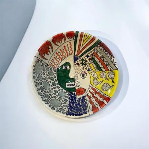 Peut inclure: Une assiette en céramique peinte à la main avec un motif de visage divisé. Un côté présente un visage vert, l'autre un visage crème. L'assiette est décorée de motifs et de formes rouges, jaunes, bleus et gris.