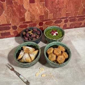 Peut inclure: Quatre petits bols texturés de couleur sarcelle remplis de divers aliments. L'un contient des olives, un autre une trempette verte avec une touche de blanc, un troisième des pâtisseries et le dernier des sucreries au sésame. Une fourchette décorative est au premier plan.