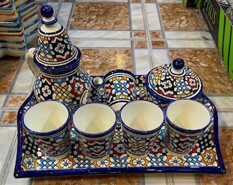 Może przedstawiać: Ceramiczny zestaw do herbaty z czajniczkiem, cukiernicą, śmietanką i czterema filiżankami, wszystkie zdobione geometrycznym wzorem w kolorze niebieskim, czerwonym, żółtym i białym. Zestaw stoi na pasującym prostokątnym tacy.