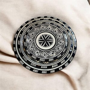 Puede incluir: Un plato decorativo en blanco y negro con un diseño geométrico. El plato presenta un motivo floral central y patrones intrincados alrededor de los bordes. El plato es redondo y parece ser de cerámica.