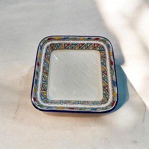 May include: Square ceramic dish with a white interior and a colorful geometric border. The border features a repeating pattern of triangles in red, yellow, green, and blue. The dish has a dark blue rim.