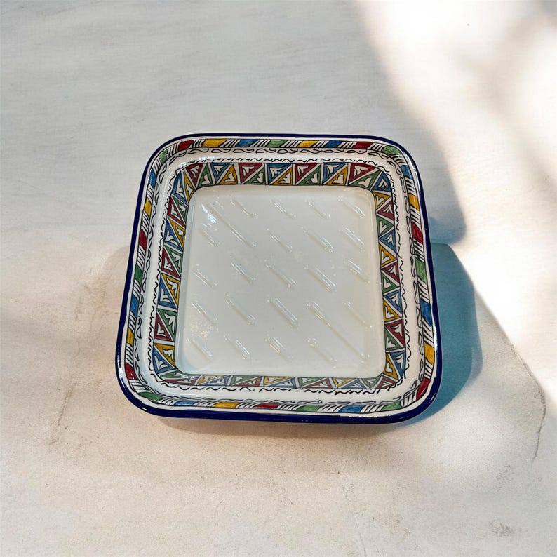 May include: Square ceramic dish with a white interior and a colorful geometric border. The border features a repeating pattern of triangles in red, yellow, green, and blue. The dish has a dark blue rim.