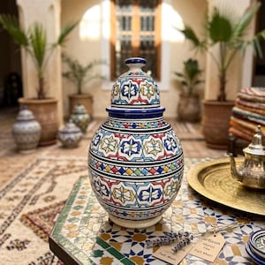 Peut inclure: Un pot en céramique avec couvercle, orné d'un motif géométrique vibrant bleu, rouge, jaune et vert. Le pot est posé sur une table en mosaïque, avec un plateau en laiton et une théière en arrière-plan. Le pot mesure environ 30 cm de haut.