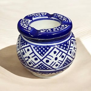 May include: A small, round ashtray with a blue and white geometric pattern. The ashtray has a circular opening with a lip and a small indentation for resting cigarettes. The ceramic ashtray is a decorative item.