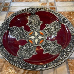 May include: A circular decorative plate with a deep burgundy centre and a silver-coloured border. The plate features a central design with a star and hand-shaped motifs. The border has an intricate, repeating pattern. The plate is likely made of ceramic or metal.