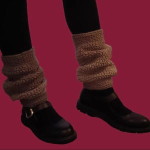 Puede incluir: Calentadores de piernas de ganchillo beige, usados sobre medias negras, con zapatos negros. Los calentadores son gruesos y texturizados, con un aspecto arrugado. Los zapatos tienen una hebilla.