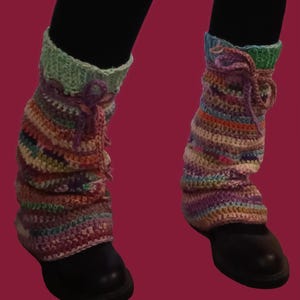 Puede incluir: Calentadores de piernas de ganchillo multicolores, con tonos de rosa, morado, naranja y verde. Se usan sobre zapatos negros y leggings negros. La parte superior es de color verde claro y está atada con una cuerda morada.