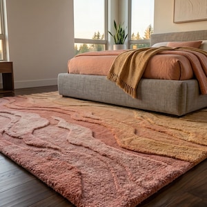May include: A large, textured area rug in shades of peach, coral, and gold. The rug features a raised, flowing design reminiscent of a landscape. The rug is placed in a bedroom setting with a bed and large windows.
