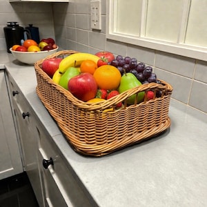 Op de afbeelding: Een rechthoekige, geweven rieten mand vol vers fruit, waaronder rode appels, groene peren, sinaasappels, druiven, aardbeien en een banaan. De mand staat op een grijze aanrecht in een keuken.