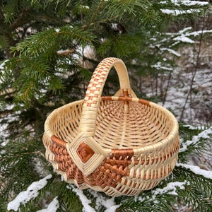 May include: A round, woven basket with a handle, featuring a natural beige and brown color scheme. The basket is decorated with diamond-shaped accents and sits among snow-covered evergreen branches.