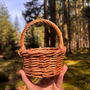 Puede incluir: Una pequeña cesta redonda de mimbre tejido con asa. La cesta es de color marrón claro y parece hecha a mano. La cesta se sostiene frente a un fondo de bosque borroso.