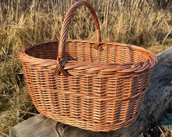 Può includere: Un cesto in vimini intrecciato con manico. Il cesto è di forma ovale ed è realizzato in vimini marrone chiaro. Il manico è anch'esso intrecciato e attaccato ai lati del cesto. Il cesto è appoggiato su un tronco di legno.