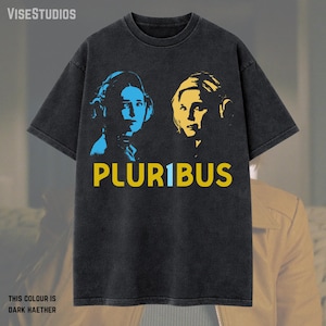 May include: Dark gray t-shirt featuring a graphic design with two faces in blue and yellow, and the word "PLUR1BUS" in yellow. The text "THIS COLOUR IS DARK HAETHER" is at the bottom. The shirt is a casual wear item.