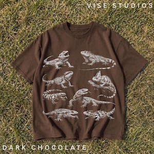 May include: A dark brown short-sleeved t-shirt featuring a white graphic print of various lizard species arranged across the front. The text VISE STUDIOS is printed in the top right corner, and DARK CHOCOLATE is printed in the bottom left corner.