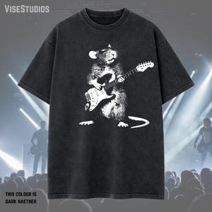 May include: Dark heather gray t-shirt featuring a white illustration of a rat playing an electric guitar. The text "THIS COLOUR IS DARK HAETHER" is at the bottom. The shirt has short sleeves and a crew neck.