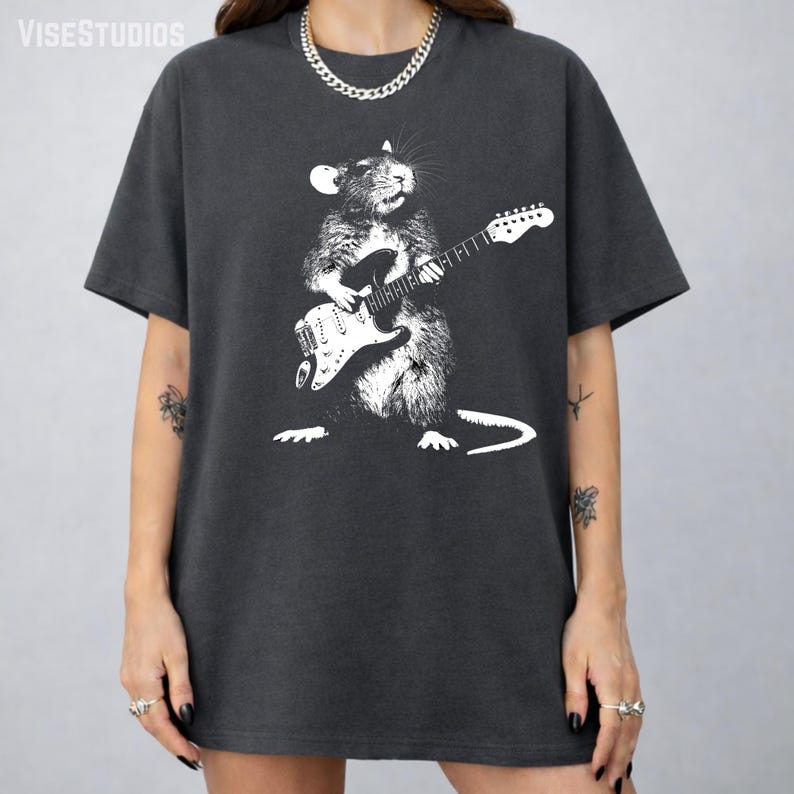 May include: Dark gray t-shirt featuring a white graphic of a rat playing an electric guitar. The rat is depicted in a detailed, artistic style. The shirt has a relaxed fit and short sleeves. The brand name "VISESTUDIOS" is printed at the top.