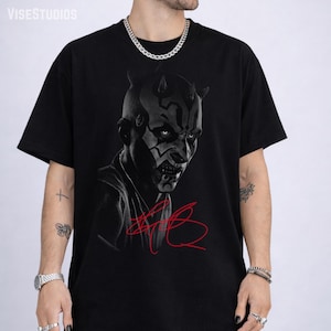May include: Black t-shirt featuring a graphic of a character with horns and facial markings. The image is in black and white with a red signature. The shirt is a crew neck.
