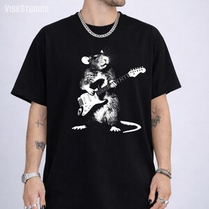 May include: Black t-shirt featuring a white graphic of a rat playing an electric guitar. The rat is depicted in a detailed, artistic style. The shirt has a classic crew neck and short sleeves. The brand name "VISESTUDIOS" is printed at the top.