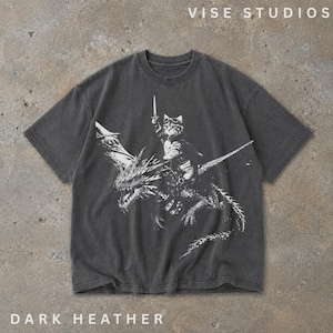 May include: A dark heather gray short-sleeve t-shirt featuring a white graphic print of a cat wearing goggles and holding a sword while riding a dragon. The text VISE STUDIOS is printed at the top right, and DARK HEATHER is printed at the bottom left.