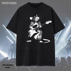 May include: Black t-shirt featuring a white graphic of a rat playing an electric guitar. The text "THIS COLOUR IS BLACK" is at the bottom. The brand name "VISESTUDIOS" is at the top. The shirt is a crew neck.
