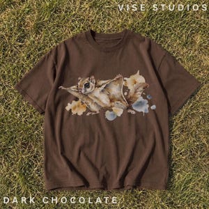 May include: A dark brown short-sleeve t-shirt featuring a watercolor-style illustration of a flying squirrel in the center. The graphic includes soft tan, brown, and blue paint splatters surrounding the squirrel. The text VISE STUDIOS is at the top.