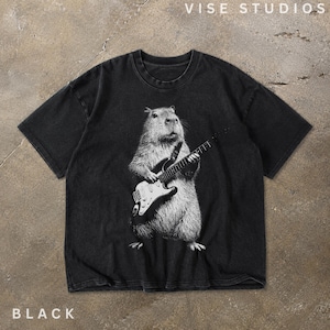 May include: A black short-sleeve t-shirt featuring a white graphic print of a capybara playing an electric guitar. The shirt has a crew neckline and a faded, washed-out appearance. The text VISE STUDIOS is printed in white at the top right corner.