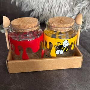 May include: Two glass jars with cork lids and wooden spoons, presented in a cardboard tray. One jar is painted red, the other yellow with a bee design. Ideal for condiments or small servings, each jar has a capacity of approximately 100 ml.