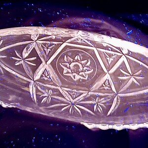 May include: Clear, oval-shaped glass serving dish with intricate starburst and geometric patterns. The dish features a scalloped edge and a central star design. The glass has a reflective quality, and the background is dark.