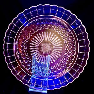 May include: A clear, vintage glass cake plate with a scalloped edge and a sunburst design in the center. The plate is supported by a clear plastic stand. The plate has a circular pattern of small circles around the sunburst.