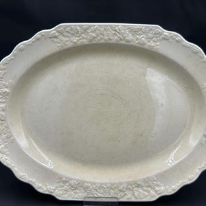 May include: Off-white oval serving platter with a scalloped edge and floral relief detailing. The platter has a slightly cracked surface and is suitable for serving food. The overall design is classic and elegant.