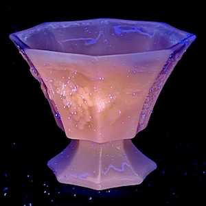 Könnte beinhalten: Eine sechseckige Glasschale mit Sockel, blau beleuchtet vor schwarzem Hintergrund. Die Schale hat einen hellrosa Farbton und eine strukturierte Oberfläche. Das Design ist geometrisch und elegant, erinnert an Vintage-Glaswaren.