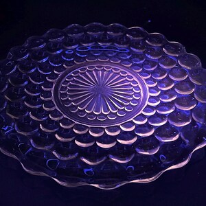 May include: A clear glass plate with a textured, bubble-like pattern around the rim and a central design. The plate has a scalloped edge and is illuminated with a purple light, creating a glowing effect.