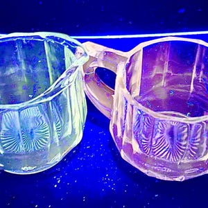 May include: Two clear, vintage glass cream jugs with handles. The jugs have a faceted design and a decorative pattern on the base. The background is a dark blue.