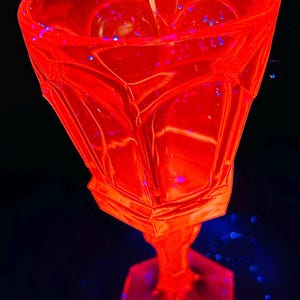 May include: A vibrant red glass goblet with a geometric design. The goblet has a hexagonal base and a detailed, angular bowl. Small blue specks are scattered around the glass, creating a striking contrast against the red. The stem is cylindrical.