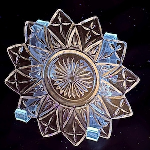 May include: A clear glass star-shaped dish with a detailed, geometric pattern. The dish has a central circular area with a sunburst design and is supported by small, clear stands. The glass has a slight iridescent quality.