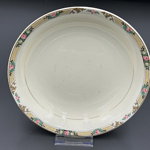 May include: A white ceramic serving bowl with a decorative floral border. The border features pink roses, green leaves, and gold accents on a tan background. The bowl has a gold rim and a simple, elegant design.