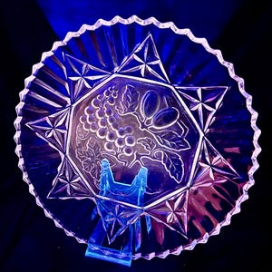 May include: Clear glass serving dish with a starburst and grape design. The dish has a scalloped edge and is supported by a blue stand. The center of the dish features a detailed grape cluster and leaf pattern.